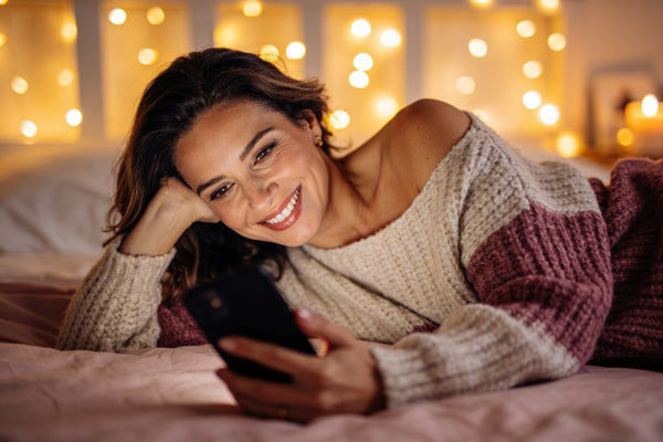 Femeie zâmbind la telefon în timpul unui apel video seara, pe pat