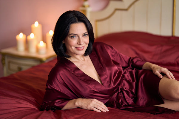Femeie elegantă în halat de satin vișiniu, relaxată pe pat cu cearșafuri roșii