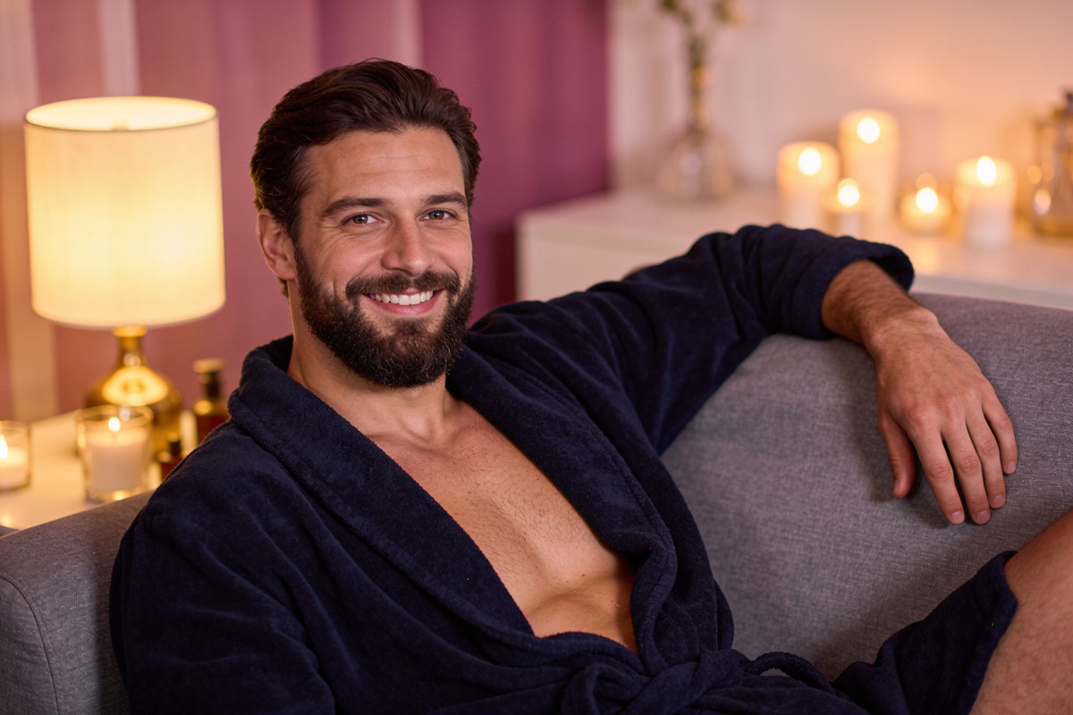 Bărbat zâmbitor în halat bleumarin, relaxat pe o canapea gri, seara