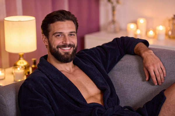 Bărbat zâmbitor în halat bleumarin, relaxat pe o canapea gri, seara
